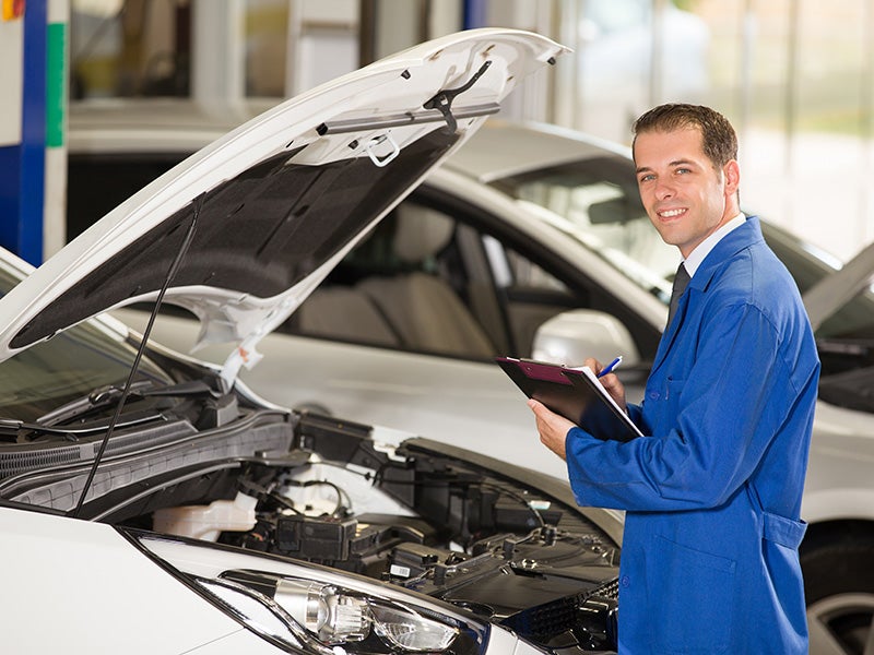 Auto technician inspecting a car engine with a clipboard in a garage.